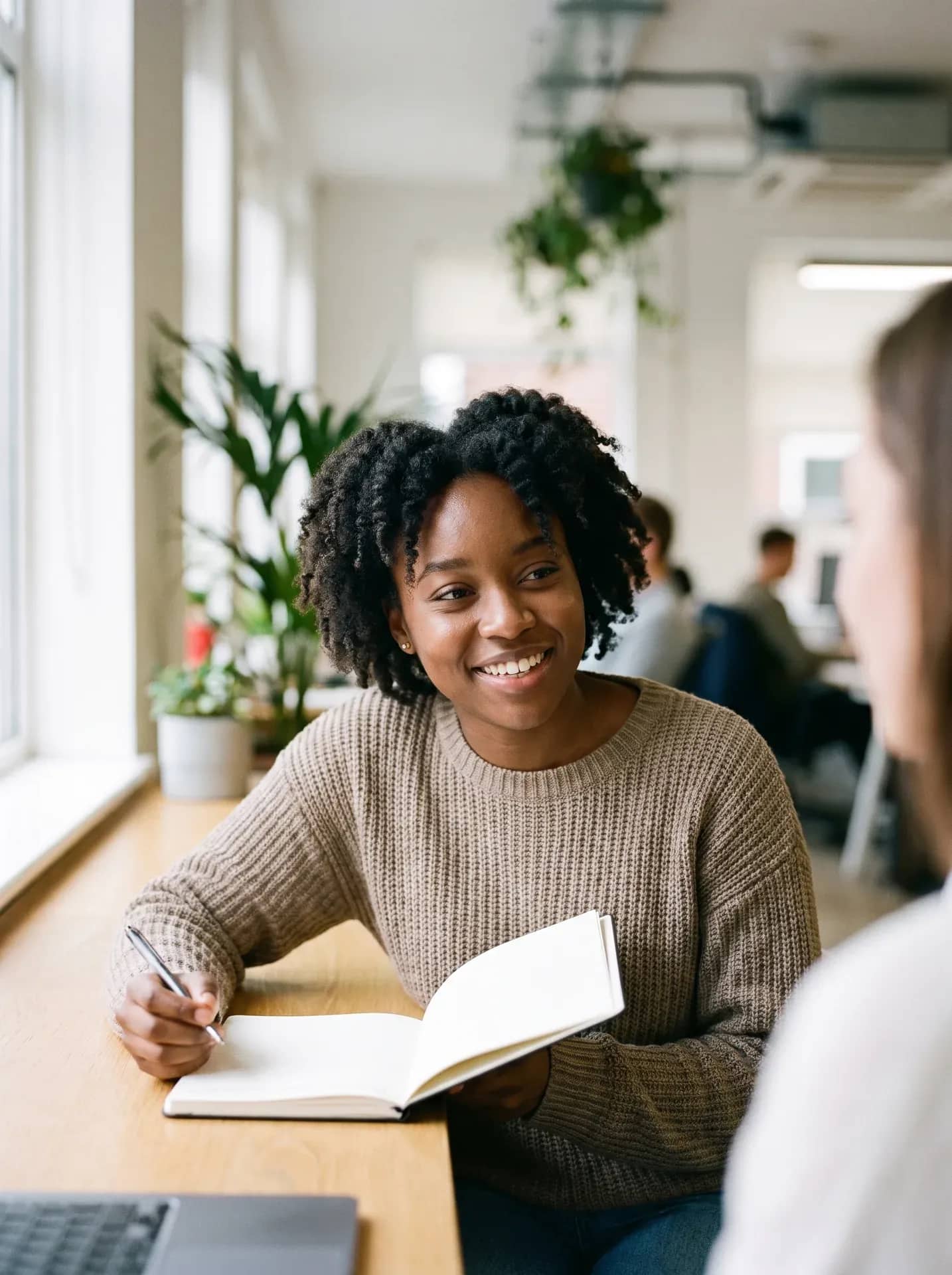 A young woman with a notebook and pen at her desk, smiling warmly at the person she is talking to just out of frame.